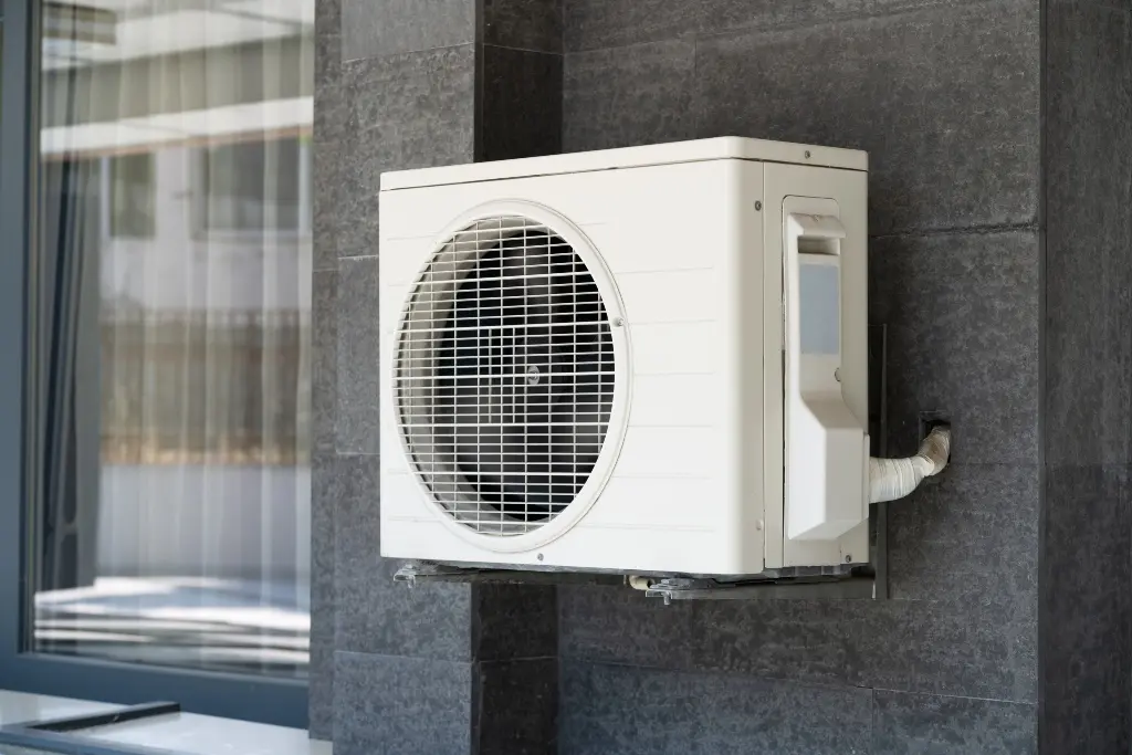White outdoor condenser unit for a mini-split air conditioning system, mounted on a dark grey tiled exterior wall.